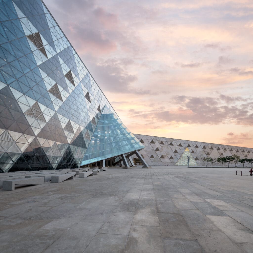 Glass pyramid structure at the Grand Egyptian Museum in Giza with triangular geometric facades against a sunset sky, featuring a large concrete plaza in the foreground